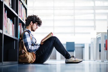 Male student in library, looking for books, preparing for final exam. Reading and sitting on floor.