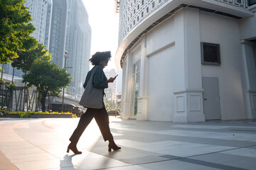 Successful African businesswoman wearing suit holding and looking at smartphone walking on street outdoors, woman wearing sunglasses and walking on street