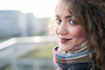 Naklejka premium Beautiful woman with curly hair standing on terrace, enjoying cold autumn morning, sunny day. Speaking positive affirmations.