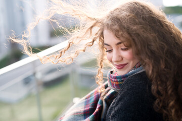 Naklejka premium Beautiful woman with curly hair standing on terrace, enjoying cold autumn morning, sunny day. Speaking positive affirmations.