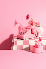 An empty pink desk decorated with a box of office supplies, behind is a table with a pink tablecloth holding a desk phone and a vase of gerbera daisy. Vacant space in center for presentation