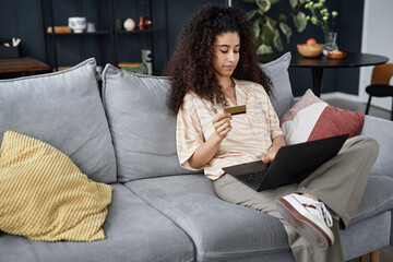 Young biracial woman sitting on couch in living room paying for goods online in modern online store...