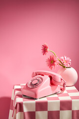 Pink background against a desk phone with pink color and a flower vase of pink gerbera daisy placed on a table has checkered pattern. Frontal shot with space for text