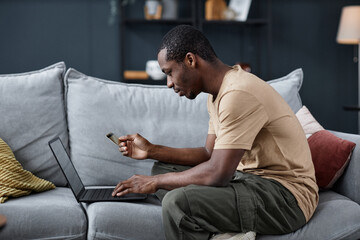 Side view of young Black man sitting on sofa in living room holding credit card browsing online...