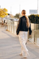 Fashionable woman with black sunglasses on her eyes and her hair in a ponytail, wearing a black leather jacket over her shoulders, white wide leg pants and white heeled boots