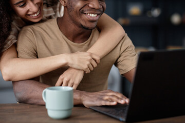 Crop shot of cheerful young biracial woman hugging her African American boyfriend while surfing Internet on laptop