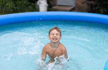 A little boy swims in an inflatable pool in the backyard of his house. A child boy happily jumps in the pool and splashes water in all directions. Summer concept, swimming pool, children.