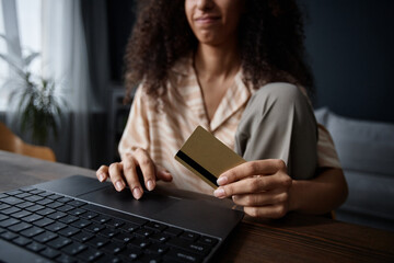 Crop shot of young biracial woman holding bank card in hand sitting at desk in living room buying goods in online store on laptop - Powered by Adobe