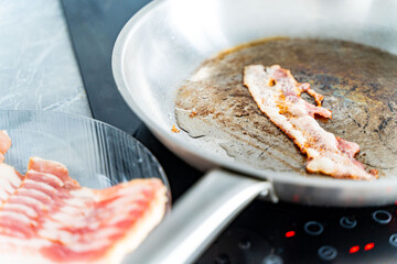 Pan-fried bacon. A strip of bacon is fried in a stainless steel pan on an induction hob. Next to a...