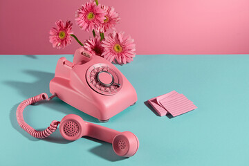 A desk phone with receiver placed on a flat blue surface, a few branches of pink gerbera flowers decorated on the back and a stack of sticky notes of the same color placed next to it. Copy space