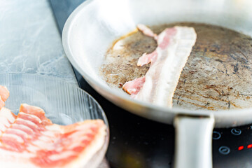 Pan-fried bacon. A strip of bacon is fried in a stainless steel pan on an induction hob. Next to a plate of raw bacon.