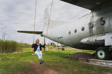 A blonde girl rides on a swing that is attached to the wing of an old An 12 cargo plane.