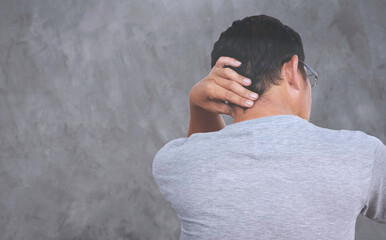 Asian middle aged man is massaging his nape because neck pain with blurred gray cement wall background, health problem concept, rear view with copy space