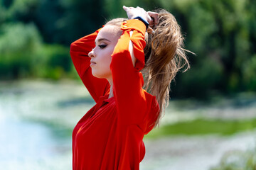 A woman in a red shirt is standing by a body of water. She has her hair in a ponytail and is...