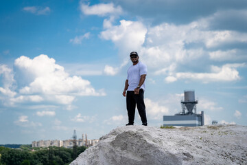 A man stands on a hill with a cloudy sky in the background. He is wearing a white shirt and black...