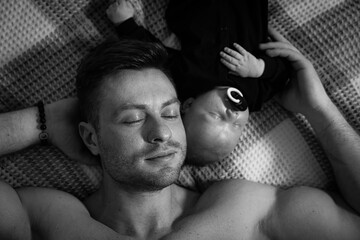 A man is laying on a bed with a baby on his chest. The man is wearing a watch and the baby is...