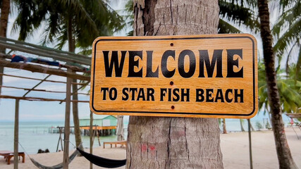 Empty yellow sign welcome hanging on a tropical tree with a serene beach, traditional boat, and clear sea in the background, ideal for summer vacation concepts