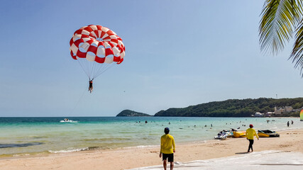 Person parasailing with a red and white parachute over a tropical beach, with onlookers and jet...