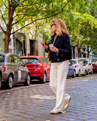 Mid aged woman holding a paper coffee cup and her mobile phone in her hand and walking down the street