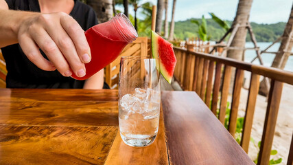 Person pouring watermelon juice into a glass at a tropical beach resort for a refreshing summer drink concept, potentially related to summer vacations or beach parties
