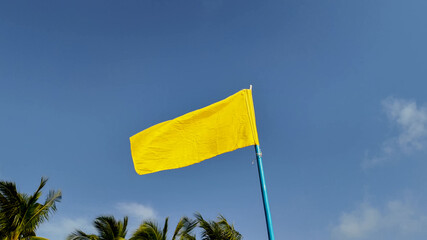Bright yellow flag fluttering against a clear blue sky with tropical palm leaves, symbolizing caution at the beach, possibly related to water safety awareness