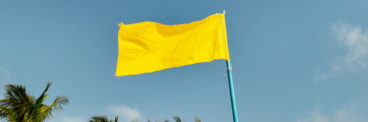 Bright yellow flag fluttering against a clear blue sky with tropical palm leaves, symbolizing caution at the beach, possibly related to water safety awareness