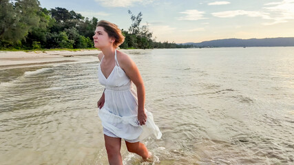 Young woman in a white dress wading in shallow beach waters at sunset, expressing carefree summer...