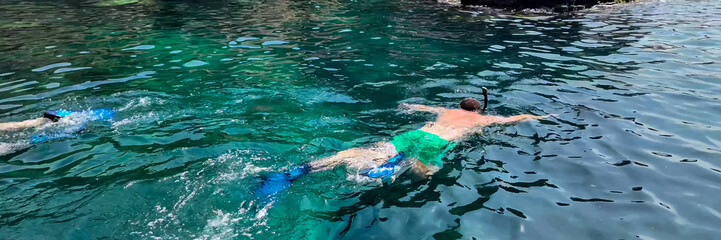 Two people swimming with snorkels in clear turquoise waters near rocky cliffs, implying summer vacations and water activities