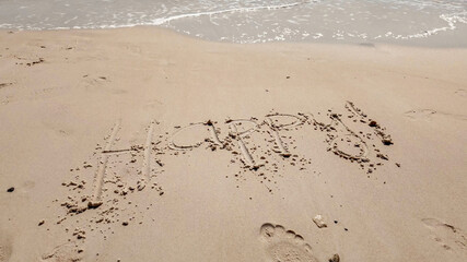 Handwritten 'Happy' message on a sandy beach with approaching waves, ideal for themes of joy,...