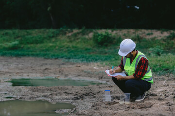 A man in red shirt and green vest is kneeling down and looking at a body of water.