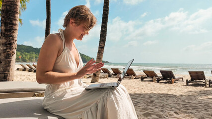 Caucasian woman using a laptop on a tropical beach with palm trees, embodying leisure technology...