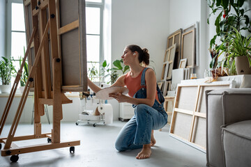 Calm female artist sits on knee holds palette brush works out small details on huge painting creates piece of art. Peaceful woman listening to music in wireless headphones focusing on creative process