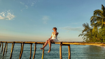 Joyful Caucasian woman in white dress sitting on a tropical beach at sunset, expressing happiness and freedom, related to travel and summer vacations