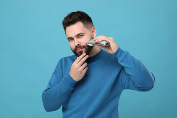 Handsome young man trimming beard on light blue background
