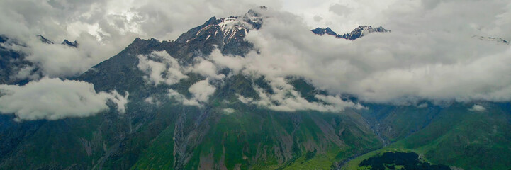 Panoramic view of majestic mountain peaks with snow caps and green valleys under a cloudy sky, ideal for nature and adventure-themed backgrounds - Powered by Adobe