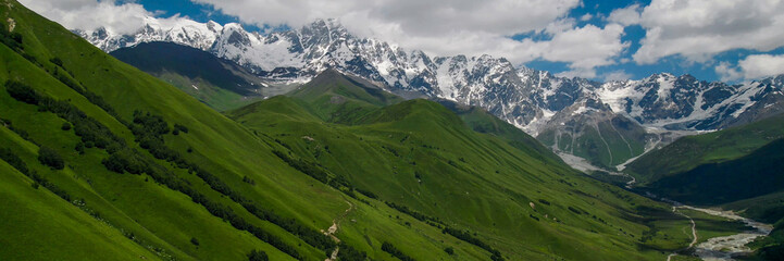 Fototapeta premium Breathtaking panoramic view of vibrant green meadows leading to snow-capped peaks under a blue sky, ideal for nature and travel themes, Earth Day promotions