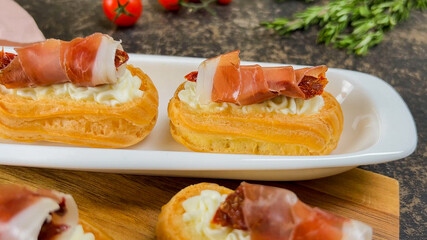Gourmet cream cheese-filled eclairs topped with prosciutto and sun-dried tomatoes on a white...