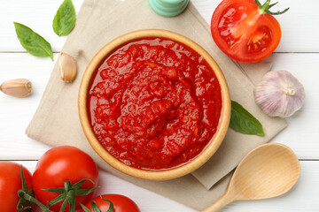 Homemade tomato sauce in bowl, spoon and fresh ingredients on white wooden table, flat lay