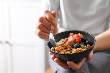 Woman eating tasty granola with berries, yogurt and seeds indoors, closeup. Space for text