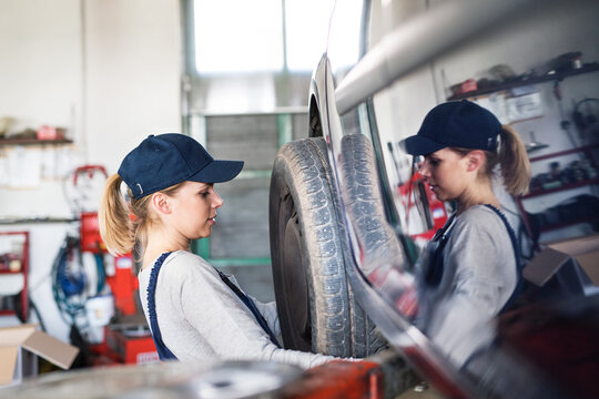 Female auto mechanic changing tieres in auto service. Beautiful woman holding tire in a garage, wearing blue coveralls. - Powered by Adobe