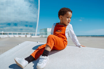 Adorable african baby child in orange overalls, sits in a ramp looking away, outdoors by the sea at...