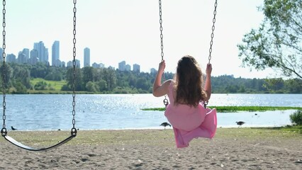 A young woman near a river lake in Canada in Vancouver girl rides swings with chains in city park smiling child in casual dress plays attraction on urban playground schoolgirl swings in sunny garden