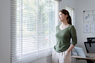  Young Asian business woman smiling confidently while standing alone in a bright modern office