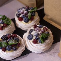 pavlova dessert with fresh blueberries, blackberries, raspberries and mint leaves on a black stone board close-up among craft coffee packs.
