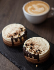 three-layer mousse dessert with chocolate and chocolate chips and crispy balls on a black stone board on a wooden table, close-up with cappuccino in a white cup on a blurred background. food photo. 