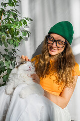 Cheerful young woman in glasses and a green beanie lovingly pets her fluffy white cat, both enjoying a sunlit cozy moment together.