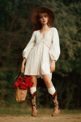A red-haired girl in a white dress and hat walks in the park with a bouquet of red tulips. Grain...