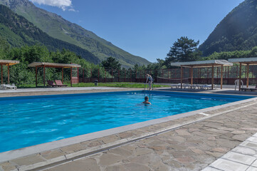 A heated pool high in the mountains. Relaxing by the water in the fresh air. Sun loungers for relaxing and sunbathing near the blue water pool. A girl and a young man are swimming in an open-air pool.