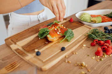 A woman hands are carefully placing slices of fresh salmon on a piece of bread on the kitchen. The sandwich is garnished with a variety of greens and seeds.
