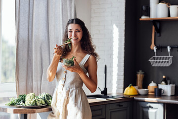 An attractive smiling girl is eating a salad of sprouted greens in the kitchen. A charming...